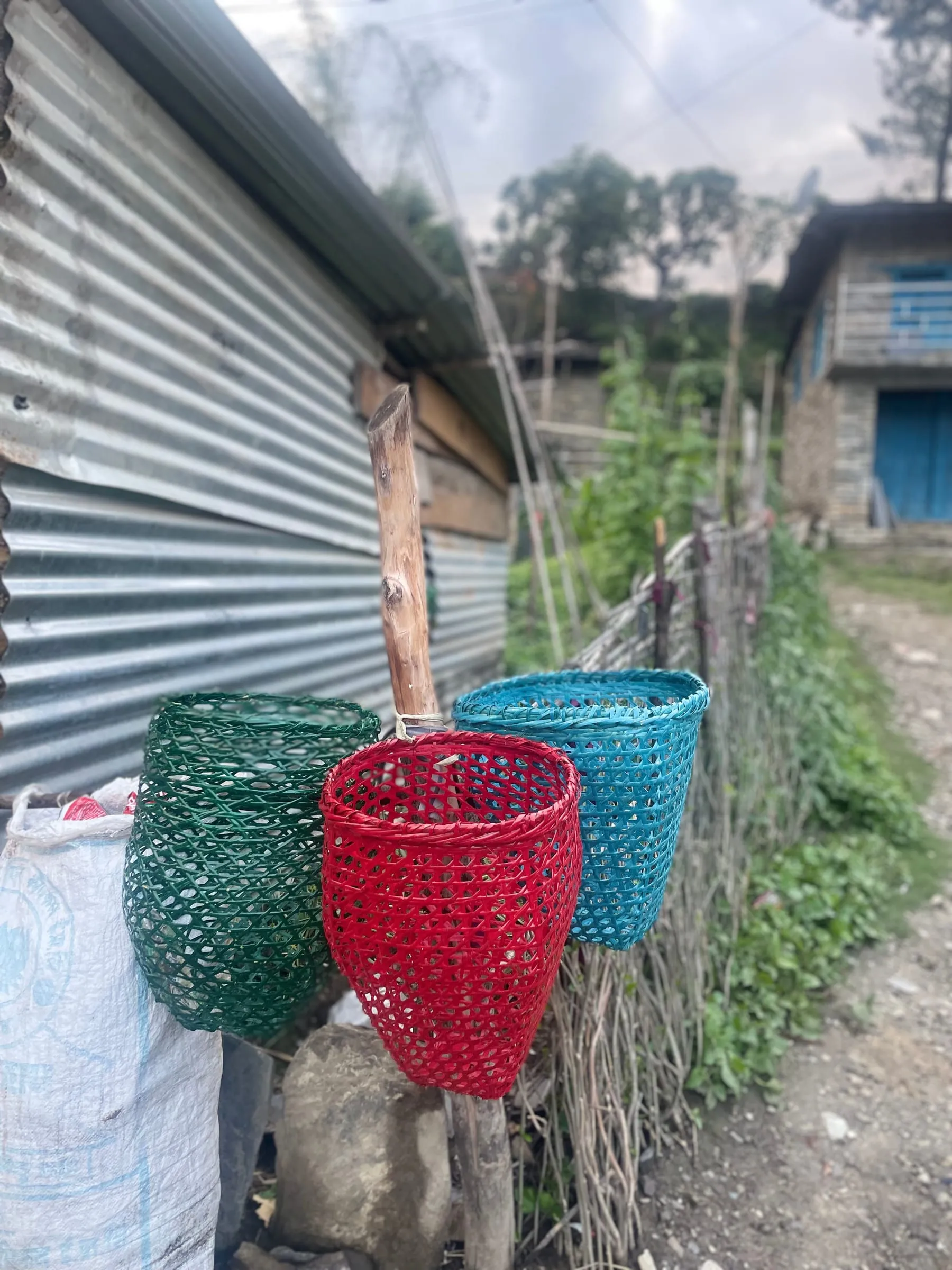 Green and red woven bamboo baskets tied to a wooden post beside a village path, sacks and a corrugated wall behind.