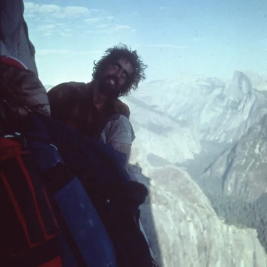 Bill Denz on a big-wall belay above Yosemite Valley, bearded and in a T-shirt, with Half Dome and the valley behind him.