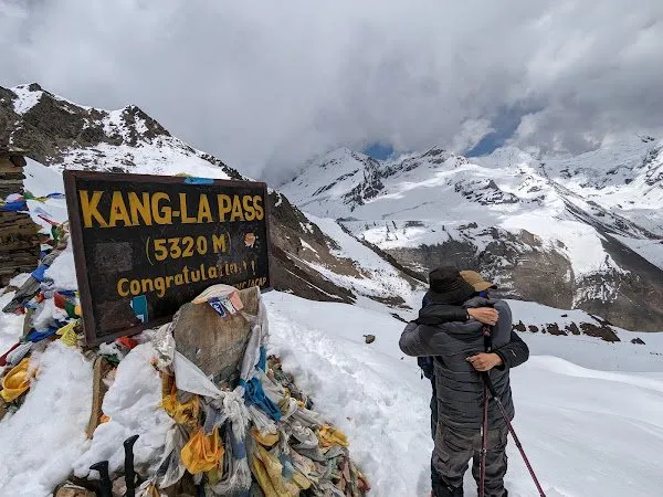 A trekker beside the Kang La Pass marker at 5,320 m, snow and cloud behind.