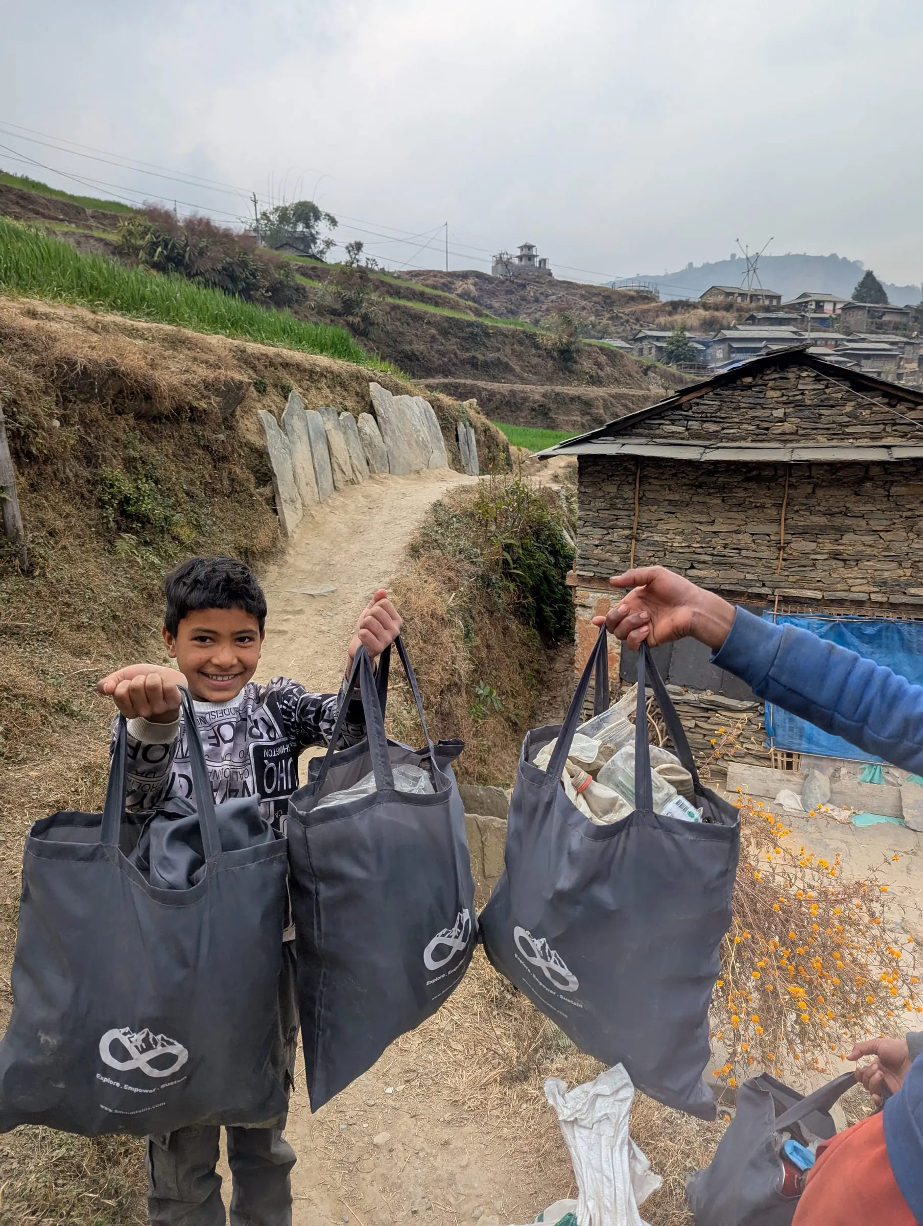 A boy on a village path holding up three 8Mountains bags filled with collected plastic, stone houses and terraced fields behind him.