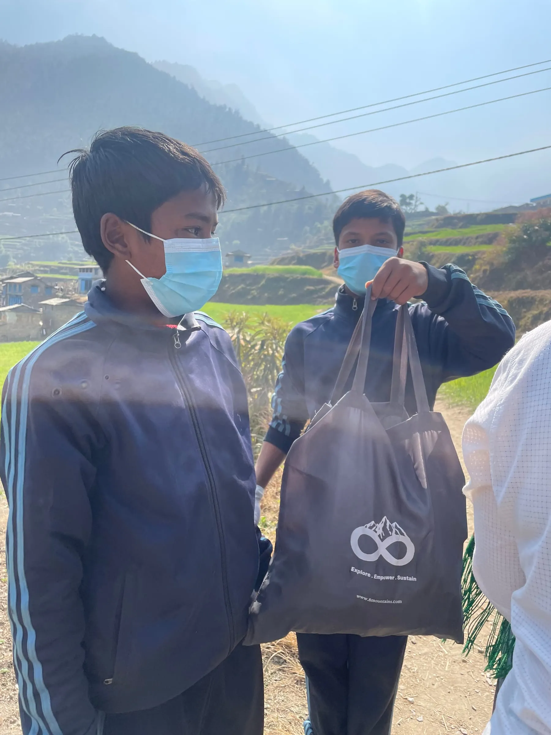 Two boys in masks holding an 8Mountains collection bag between them, terraced fields on the slope behind.