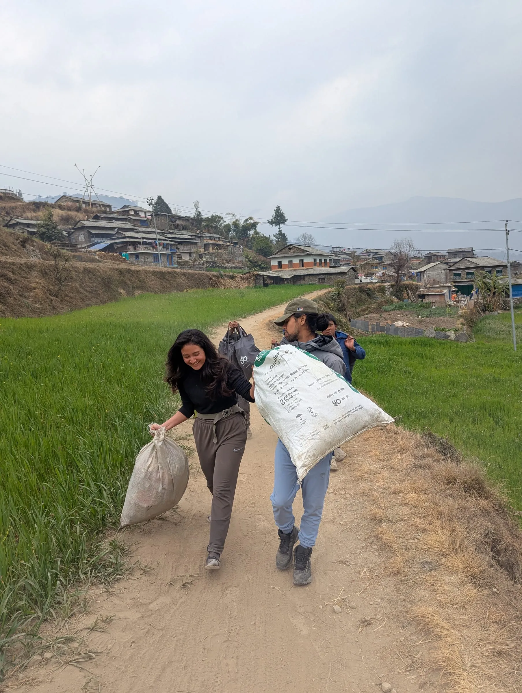 Two women hauling full sacks up a dirt path between green terraced fields, the village on the hillside beyond.