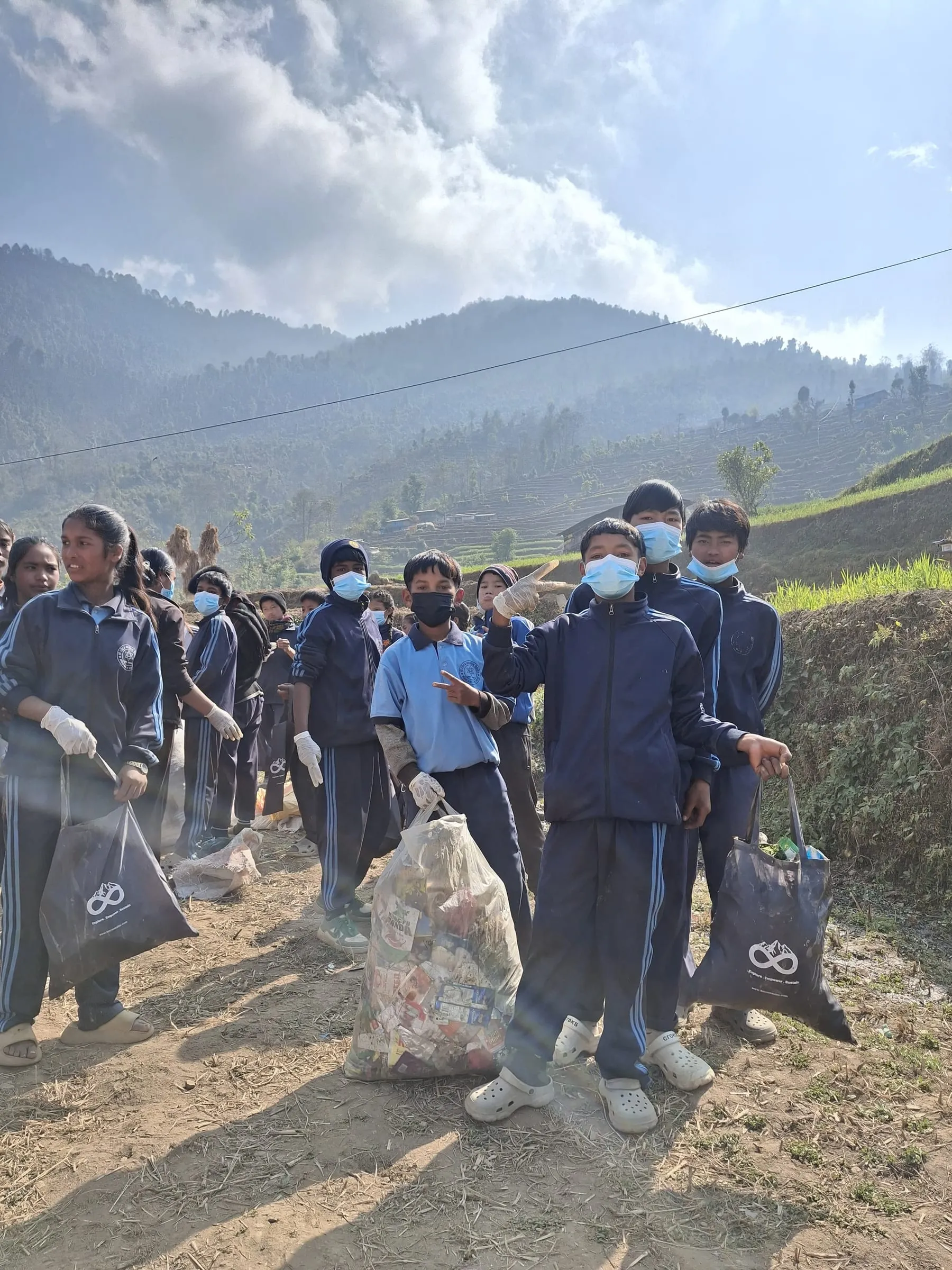 Schoolchildren in face masks and white gloves walking a hillside path, carrying clear and branded bags of collected plastic.