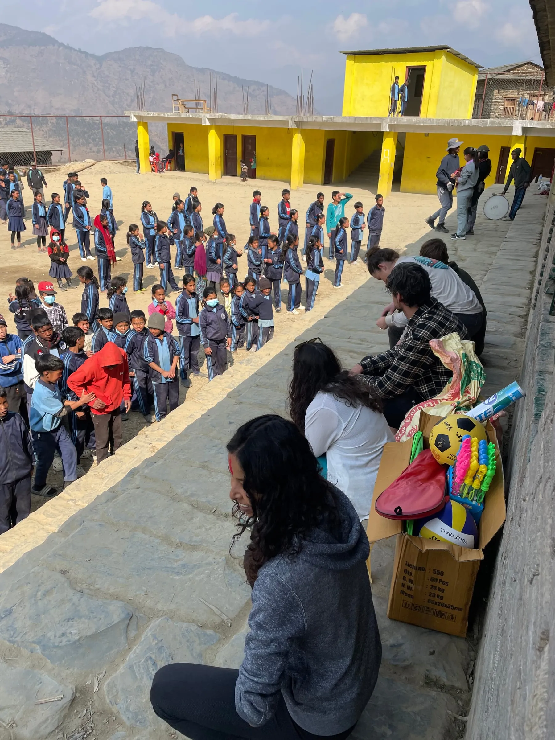 Children queuing along a wall at the school while a volunteer kneels beside boxes of prizes and sports equipment laid out on the ground.