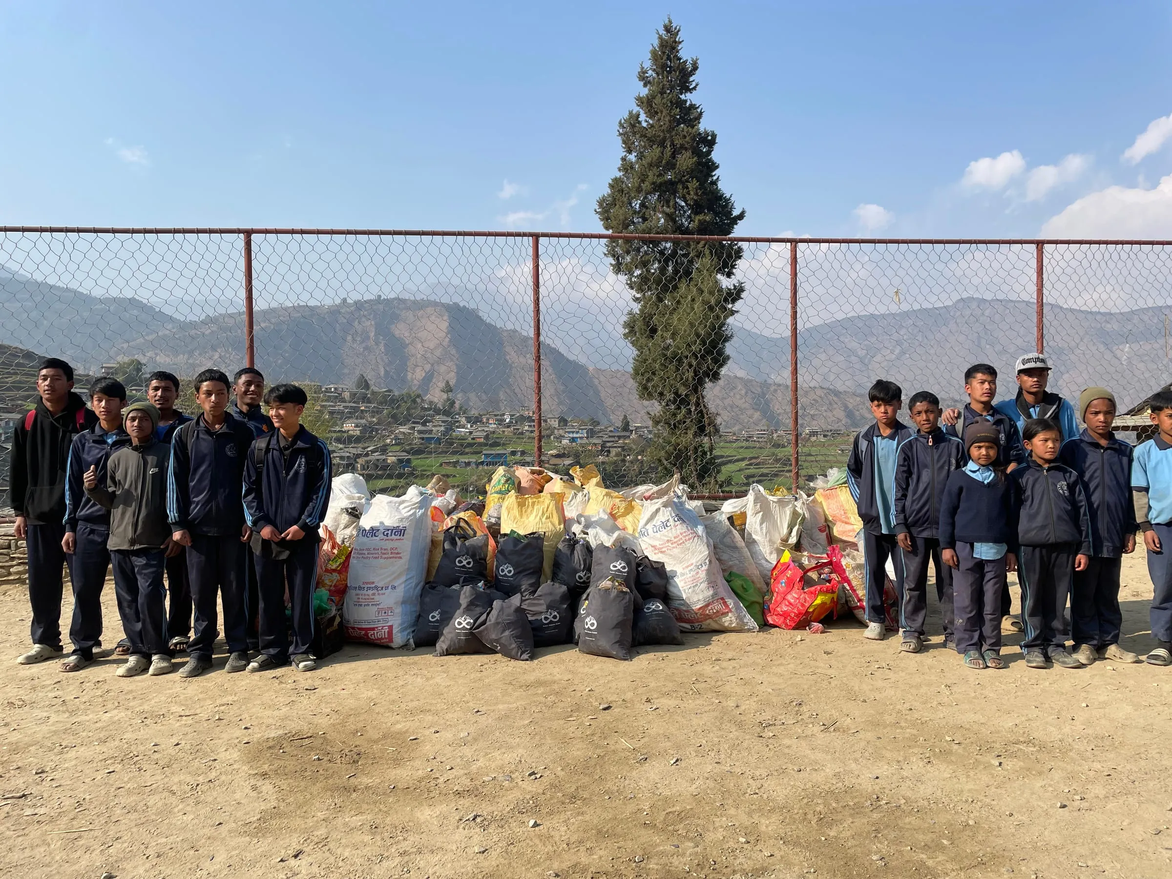 Students standing in a line behind a long row of filled sacks and bags at a wire fence, hills in the distance.