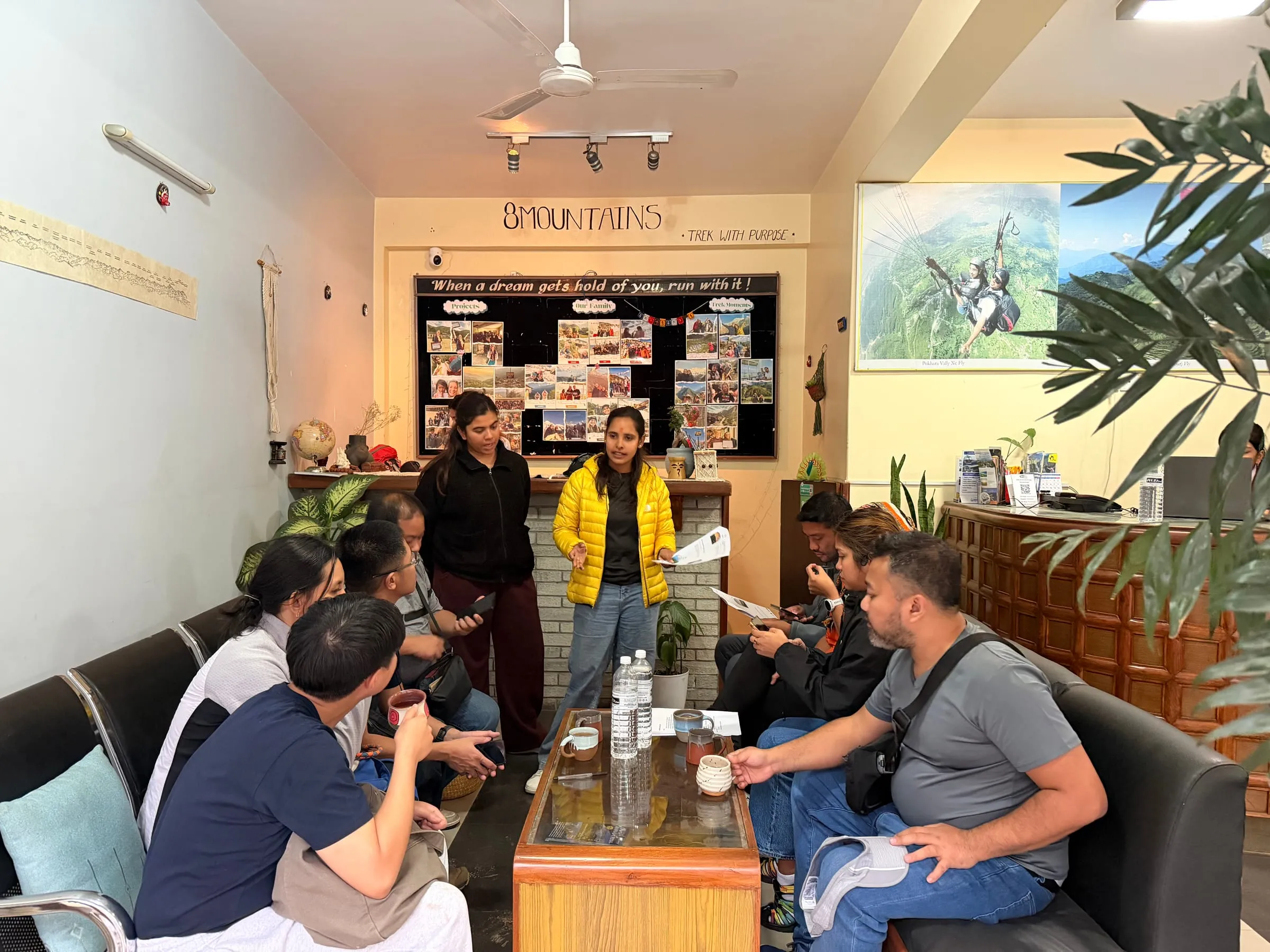 A briefing in the 8Mountains office: a guide in a yellow jacket standing and talking to a group of travellers seated around a low table, with the 8Mountains sign, a board of trek photographs and plants behind her.
