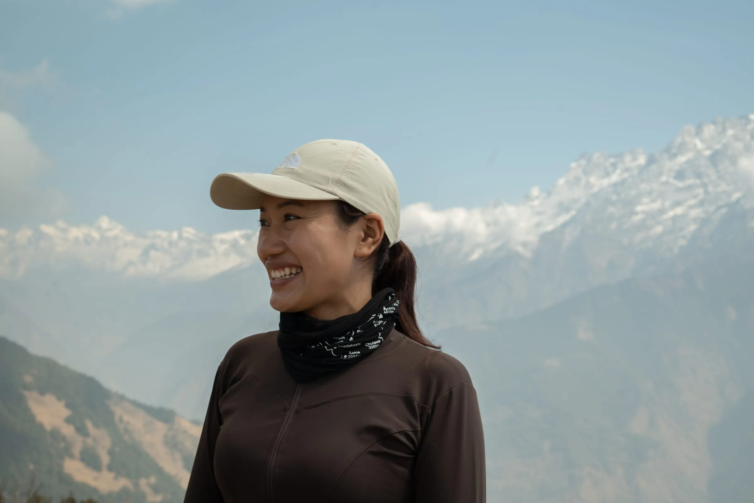 A guide in a cap and buff standing on a ridge, smiling as she looks out across a wide hazy range of foothills.