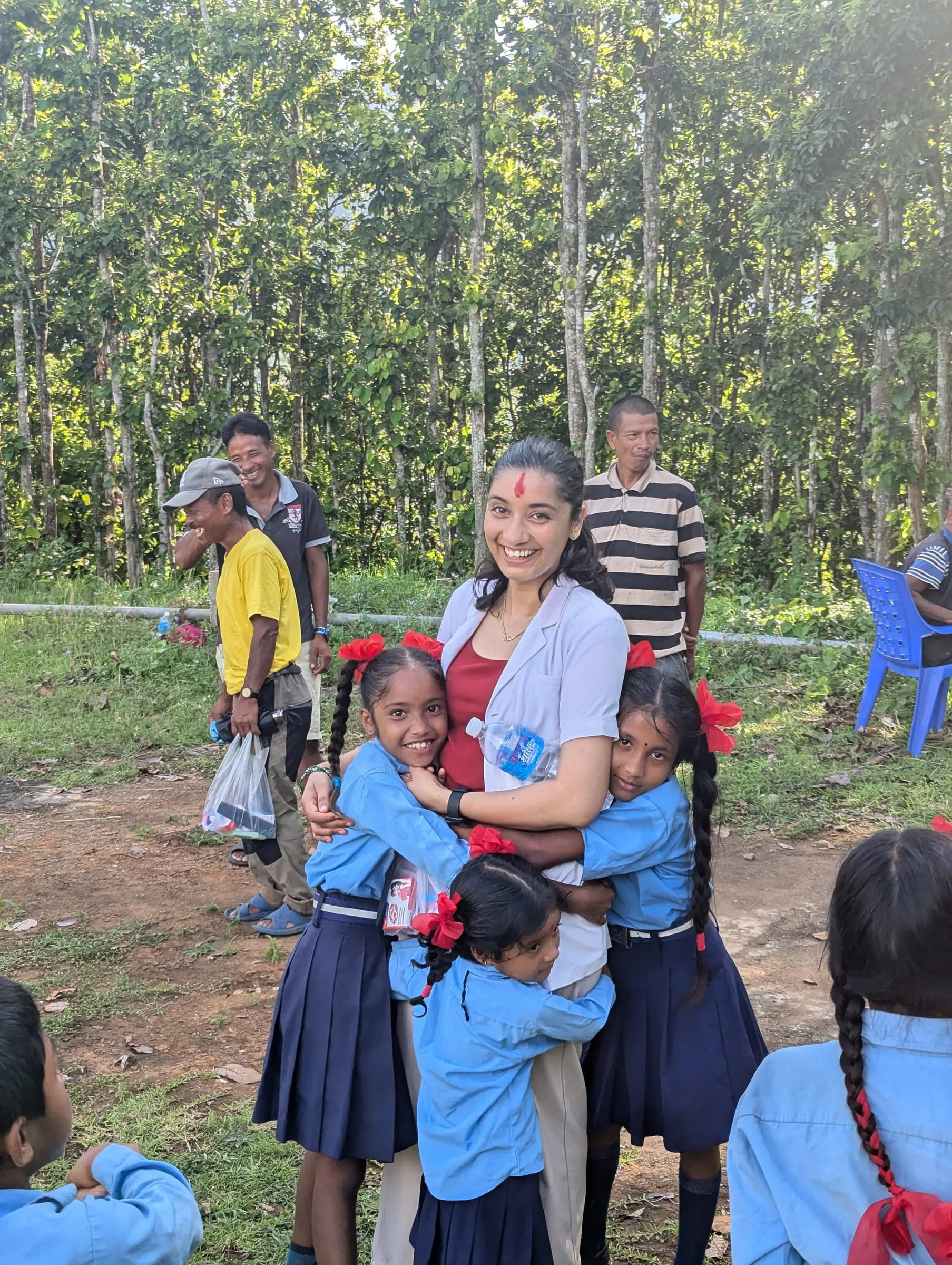 A health worker in a white coat with a tika on her forehead, being hugged by three schoolgirls in blue uniforms with red ribbons at an outdoor camp, trees and waiting families behind.