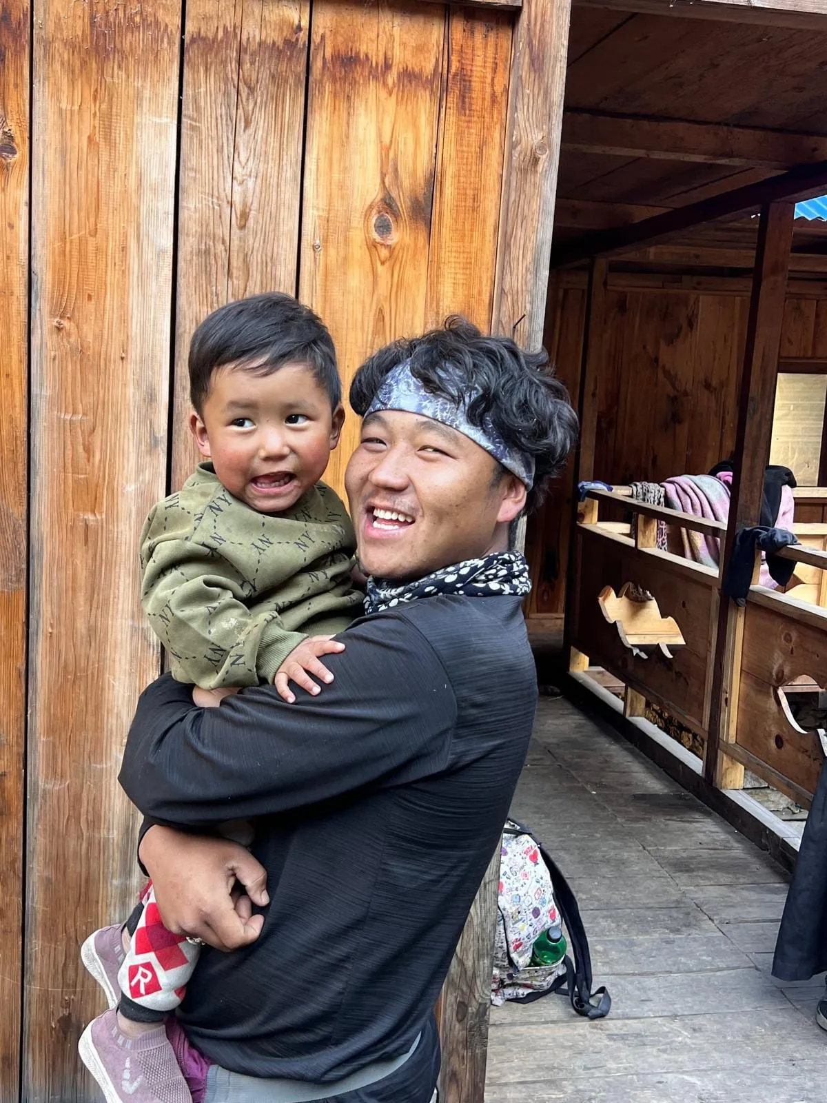A porter in a patterned headband carrying his laughing young son on one arm, on the wooden veranda of a teahouse.