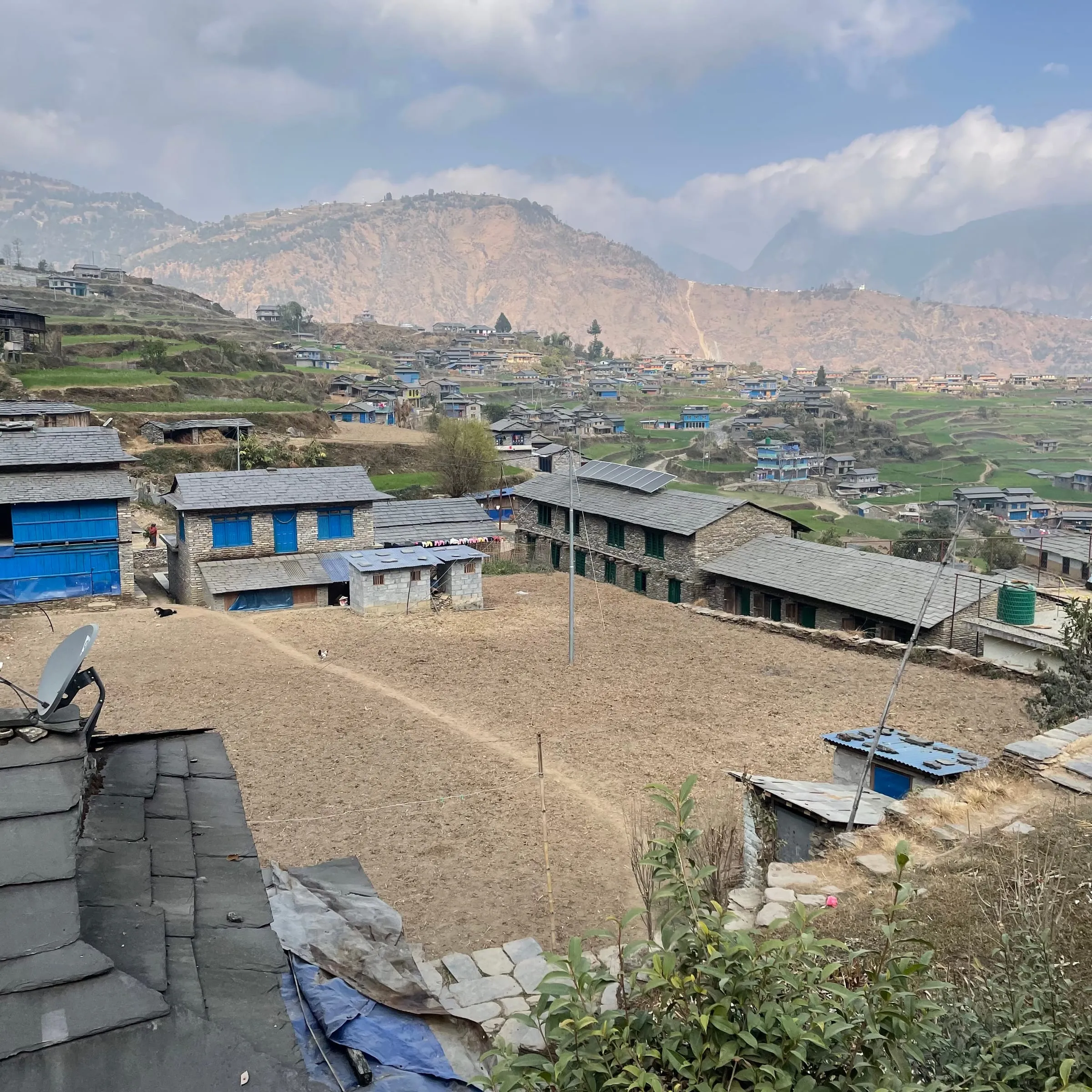 Ruma seen from a rooftop: slate-roofed stone houses around an open dusty ground, terraced fields climbing the hillside and hazy ridges behind.
