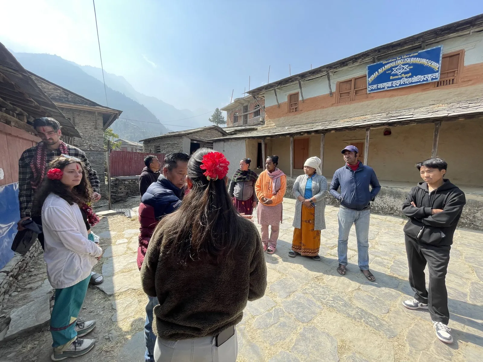 Villagers standing together in an open space between village houses, talking, hills rising behind the rooftops.