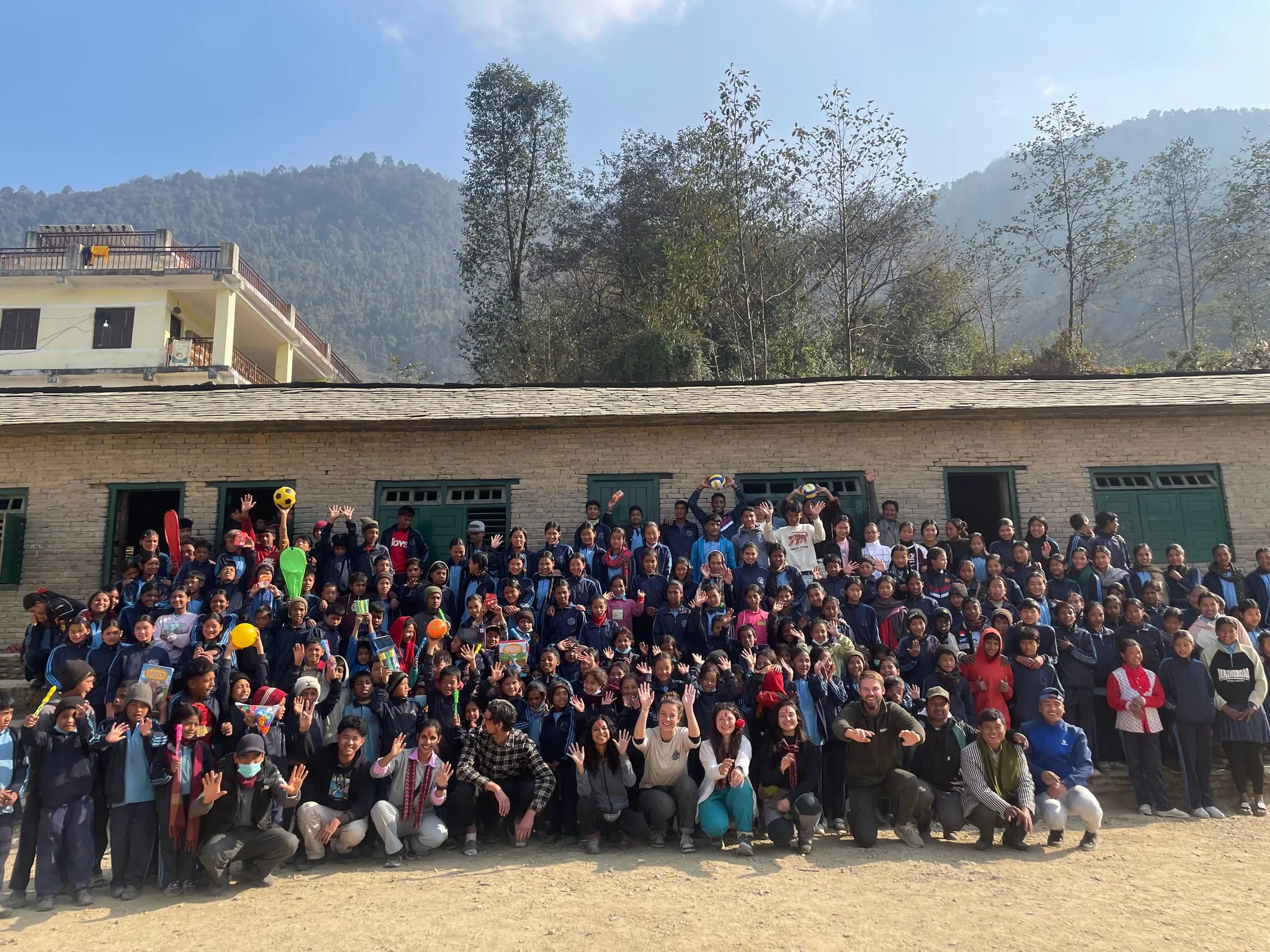 The whole school assembled in front of a long stone schoolhouse for a group photograph, many of the children with their hands in the air.