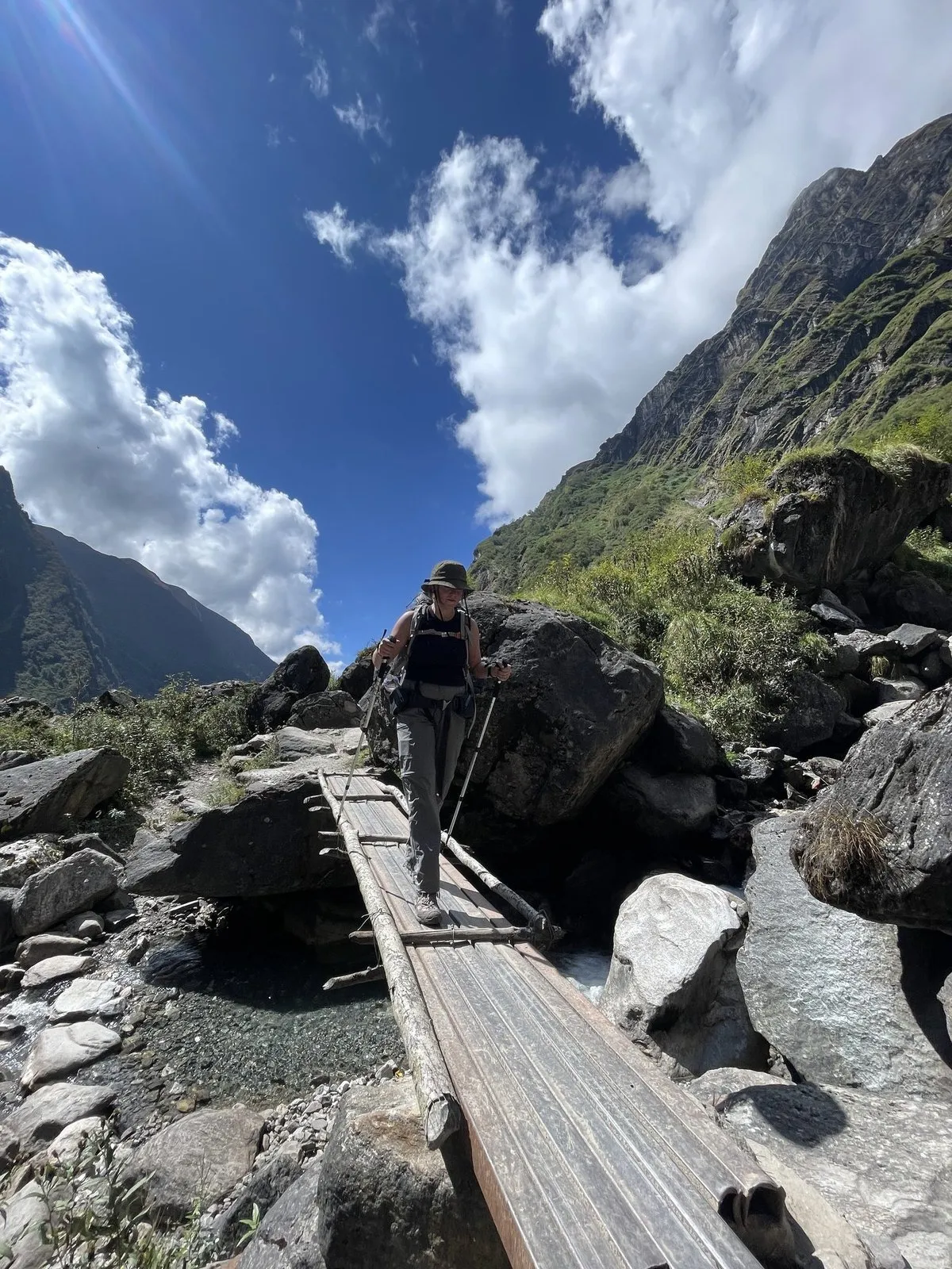 A trekker crossing a plank bridge over boulders in the Modi Khola gorge, valley walls rising steeply either side.