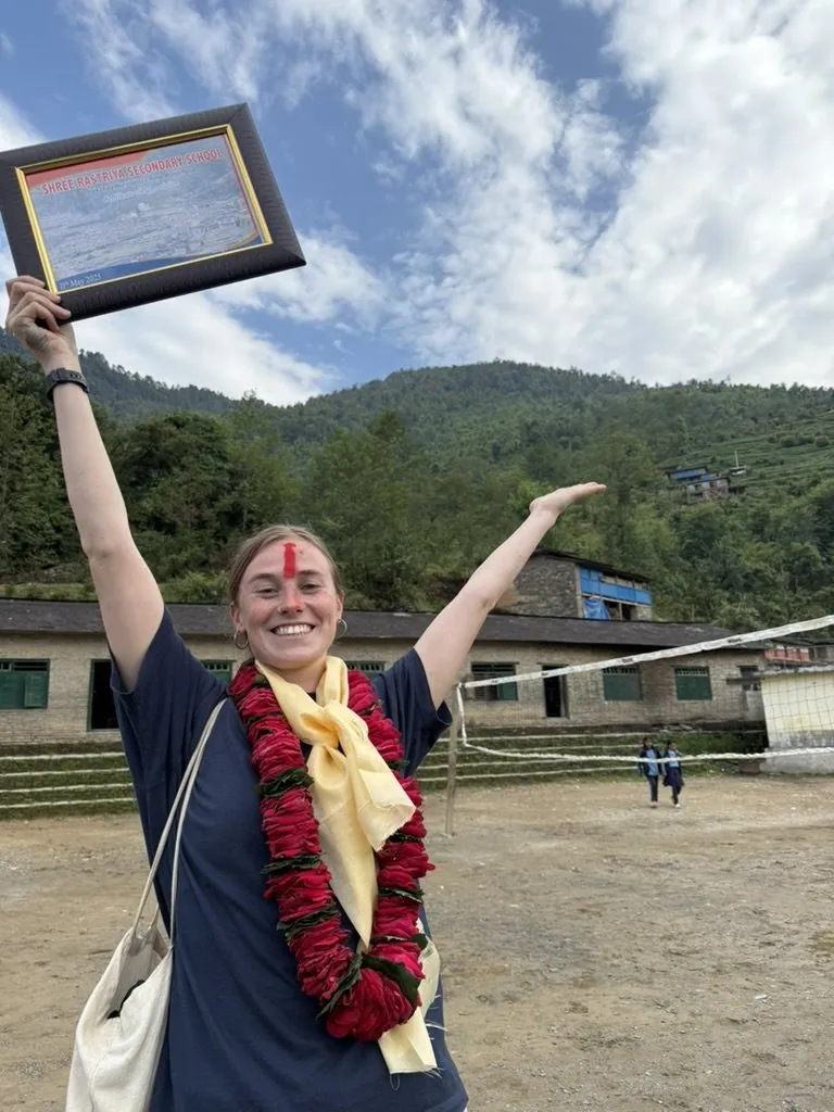 Franka, a volunteer in Ruma, wearing a garland and tika and holding up a framed certificate with both arms.