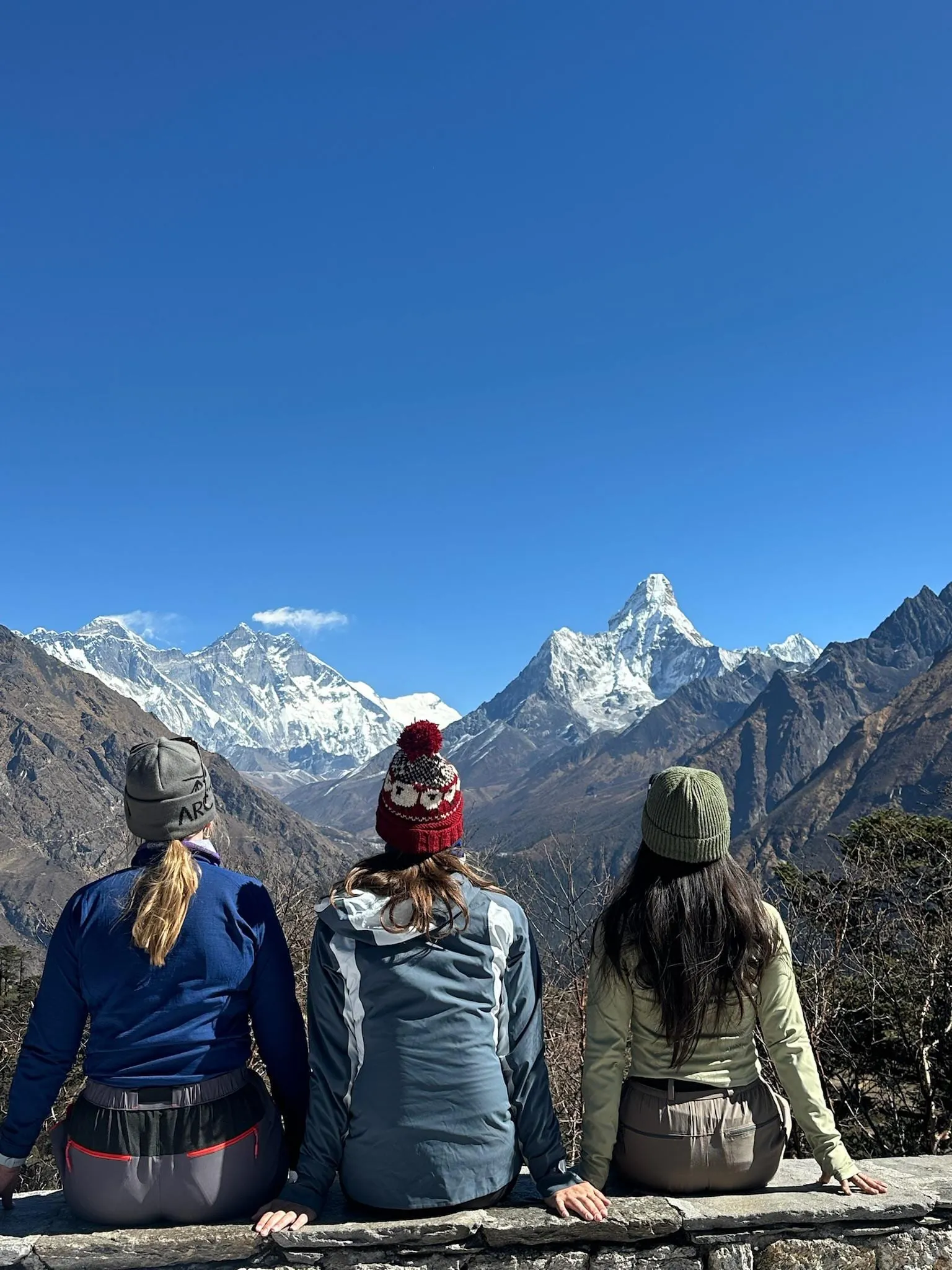 Three women sitting on a stone wall with their backs to the camera, looking out at Ama Dablam and the snow peaks of the Everest range.