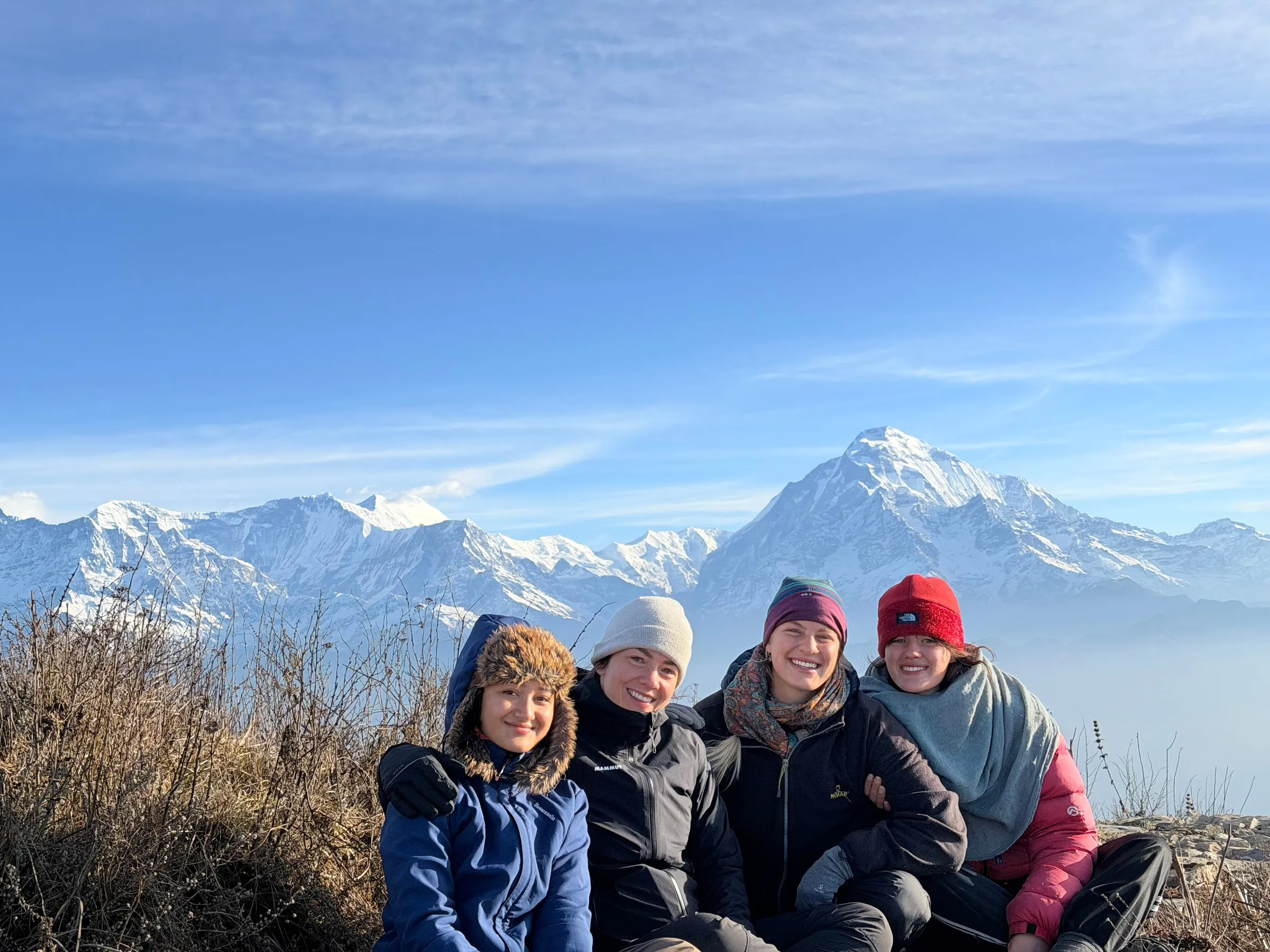 Four women sitting together on a summit at sunrise in down jackets and woollen hats, a long line of snow peaks and a sea of cloud behind them.